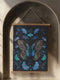Wall hanging tapestry featuring a large copper butterfly with turquoise and azure blue markings surrounded by blue ginkgo leaves on black background. The textile hangs from wooden dowels with twine, showing detailed 3D embroidery-like texture.